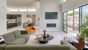 A modern living room with a sofa, coffee table, and a fireplace. There is a dining area with chairs and a table, and a kitchen in the background.