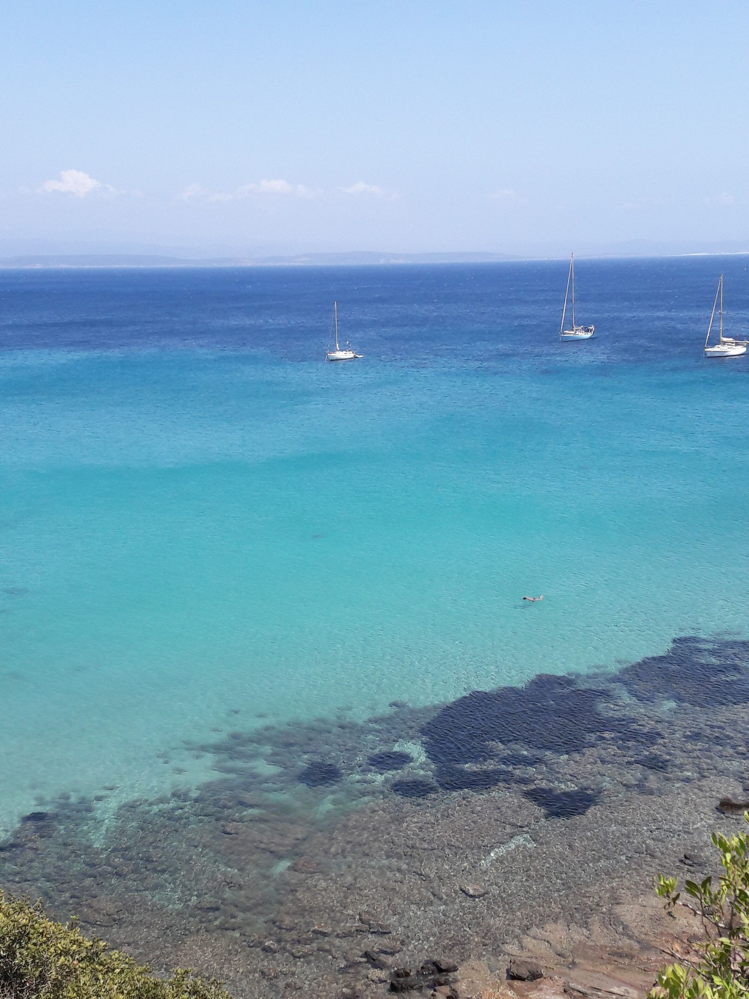 A coastal scene with clear turquoise waters, rocky shoreline, and three sailboats anchored in the distance.