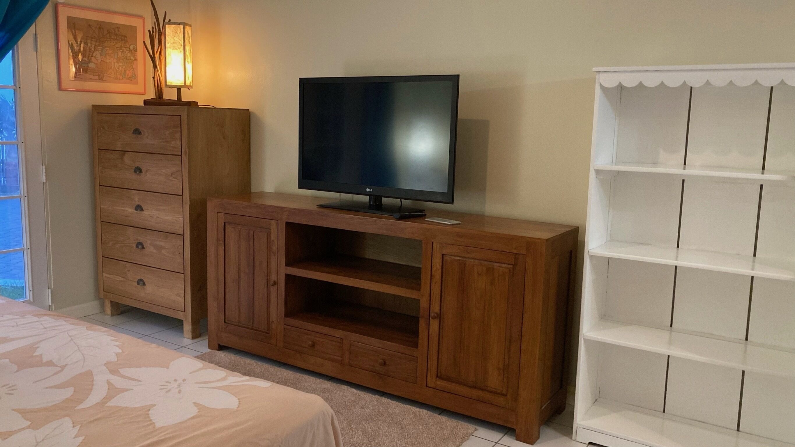 Een slaapkamer met een houten tv-meubel, een flatscreen televisie, een witte plankunit, een bed met een bloemenbedekking en een ingelijst schilderij aan de muur.