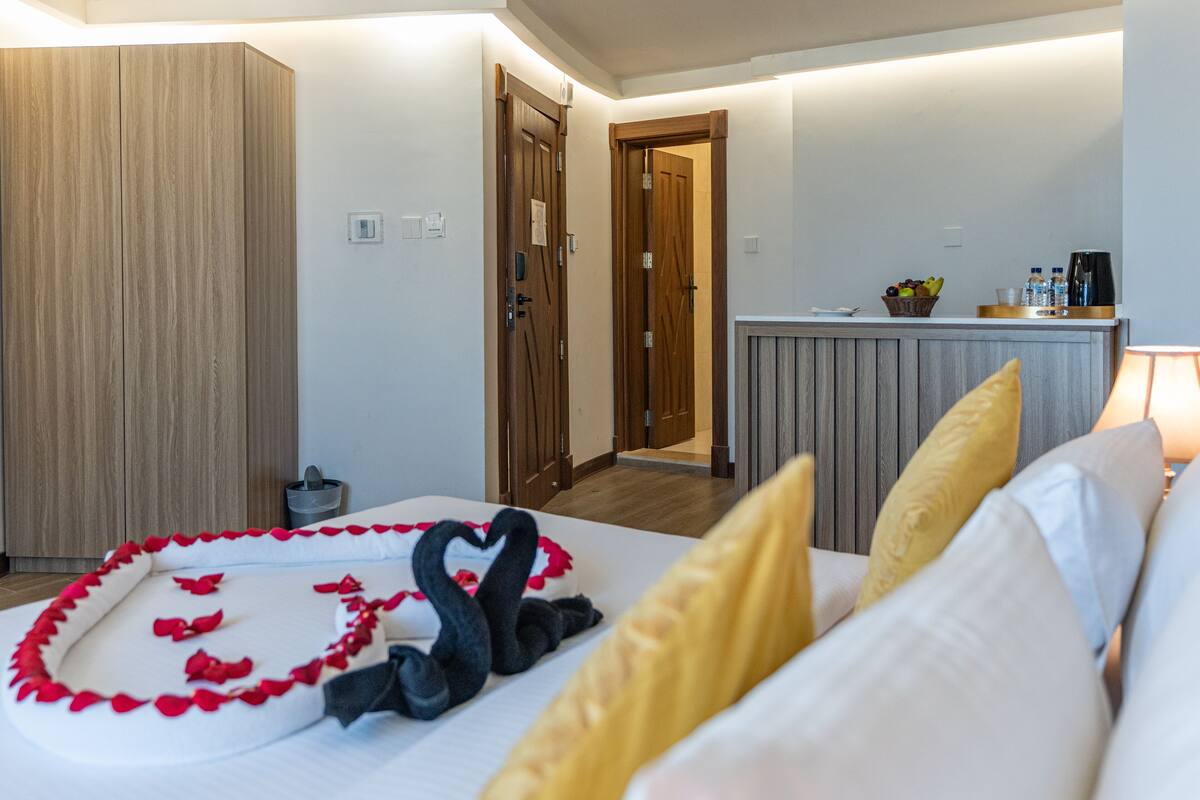 A hotel room with a bed, a wooden wardrobe, a kitchenette, and a bathroom visible through an open door.
