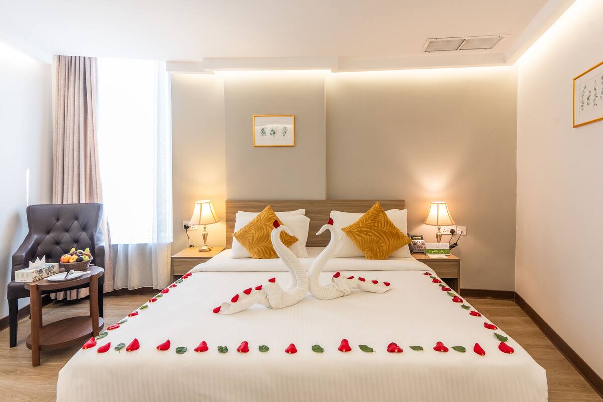 A hotel room with a neatly made bed, two lamps on either side, a chair, and a bedside table with a fruit bowl.