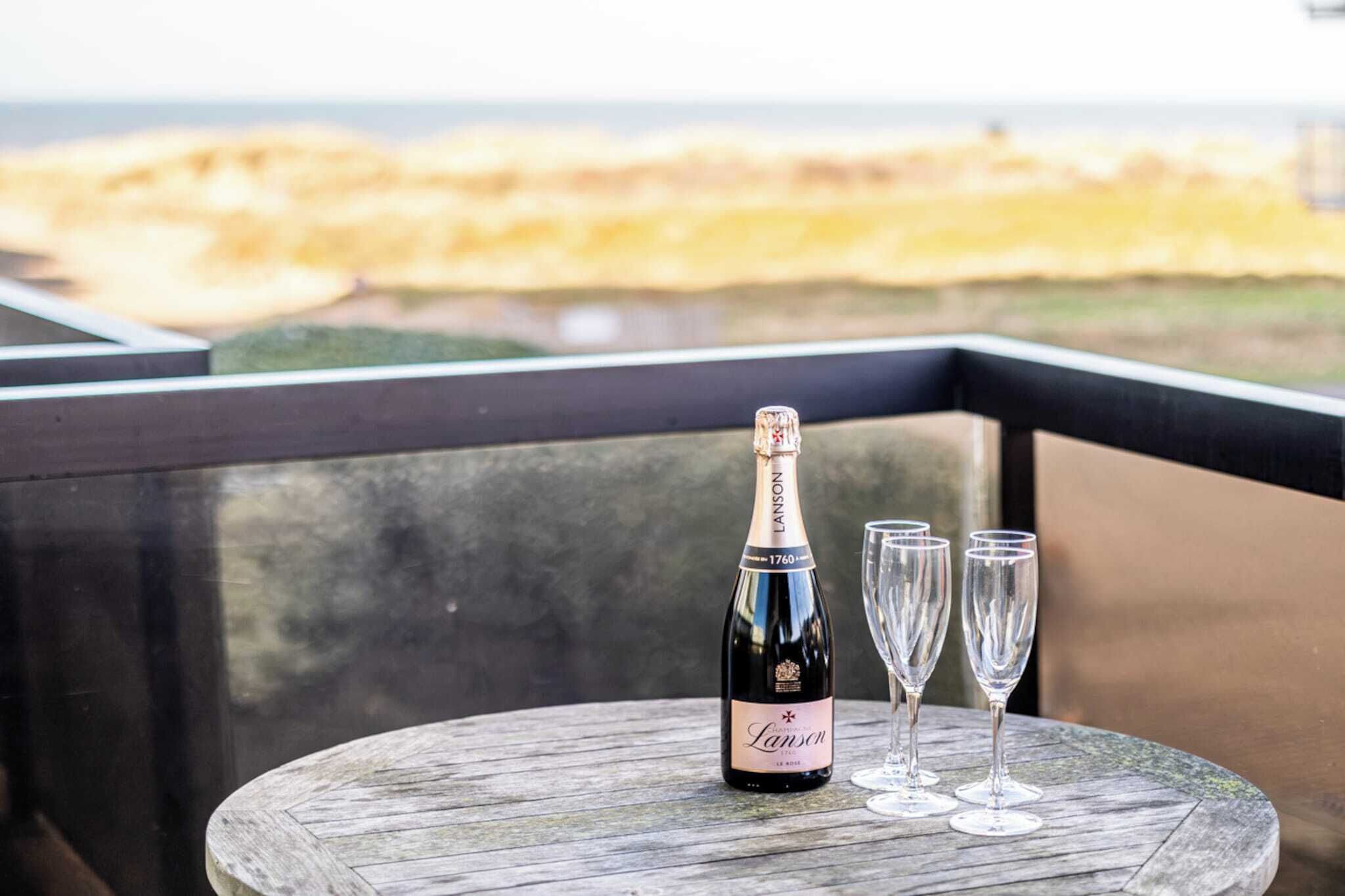 A bottle of Lanson champagne and three glasses on a wooden table with a view of the beach.