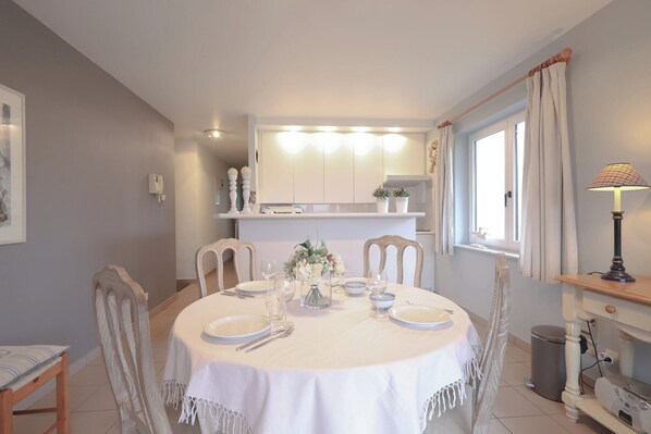 Un comedor con una mesa redonda dispuesta para cuatro personas, rodeada de sillas. Se observa una cocina al fondo con armarios y una ventana.