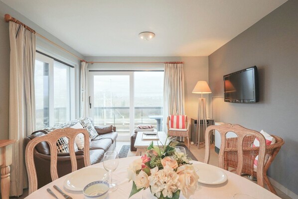 A well-lit living room with a dining table set for two, a sofa, and a television mounted on the wall.