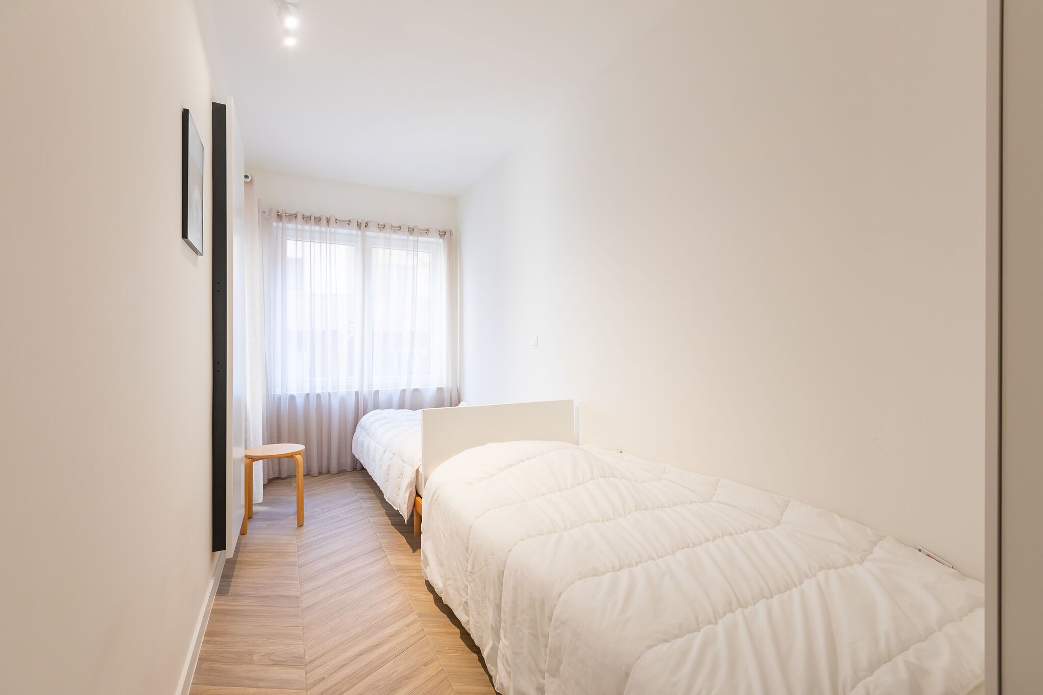 Um quarto pequeno e iluminado, com piso de madeira, uma cama de solteiro, uma mesa pequena e uma janela com cortinas leves.