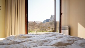 A bedroom with a large bed, a window with curtains, and a view of buildings and trees outside.