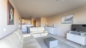 A modern living room with a white sofa, a small coffee table, a TV on a white cabinet, and a painting on the wall.