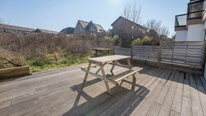 A wooden deck with a picnic table and benches, surrounded by a wooden fence and vegetation.