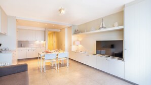 A modern kitchen with white cabinetry, a dining area with a table and chairs, and a TV mounted on the wall. - Apartment by Belgian Coast with Beach Views (Oostduinkerke)