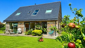 A two-story house with a sloped roof, a patio with a table and chairs, and a garden with fruit trees.