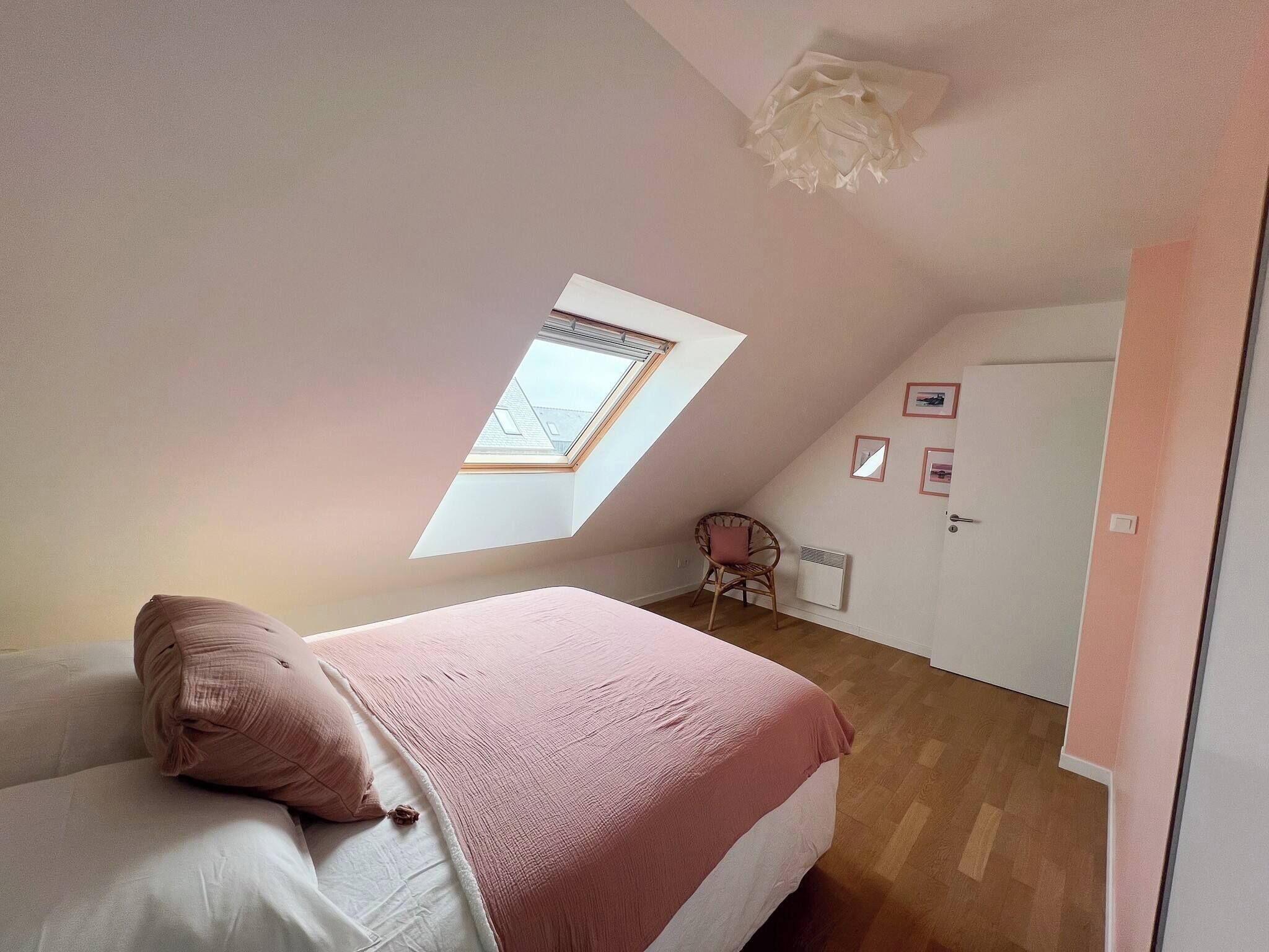 A bedroom with a sloped ceiling, a skylight, a bed with pink bedding, a chair, and a door with framed pictures.