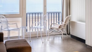 Un balcon avec des chaises en verre transparent et un fauteuil en osier, donnant sur une plage.