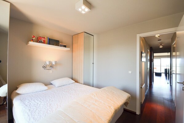 A hotel room with a large bed, a wardrobe, a shelf with books, and a hallway leading to another room.