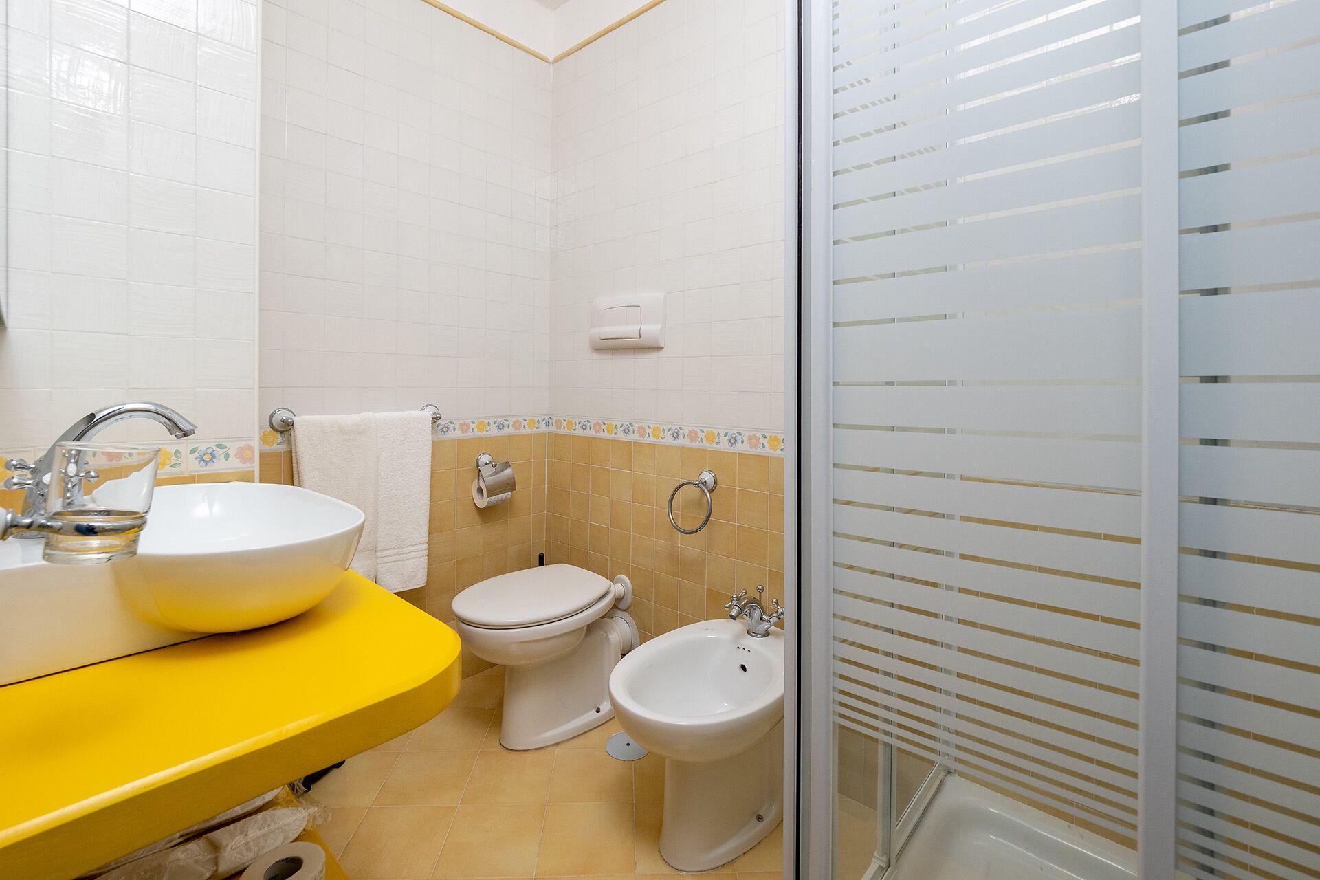 A bathroom with a yellow sink, white toilet, and bidet, with beige tiles and a glass shower enclosure.