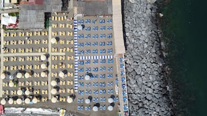 An aerial view of a beach with rows of sun loungers and umbrellas, a rocky shoreline, and a building in the background. - Marea Luxury Rooms (Vico Equense)