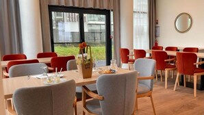 Un espace repas avec des tables et des chaises en bois, un vase rempli de fleurs et une vue sur l’extérieur grâce à de grandes fenêtres.