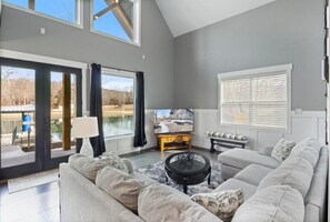 Ein modernes Wohnzimmer mit grauem Sofa, Couchtisch und Fernseher. Ein großes Fenster mit Jalousien, eine Lampe und Blick auf ein Haus im Freien.