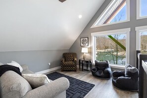 Un salón estilo loft con techo alto, un sofá gris, un sillón estampado, un asiento informal tipo puff negro, y vistas a un parque a través de grandes ventanales.