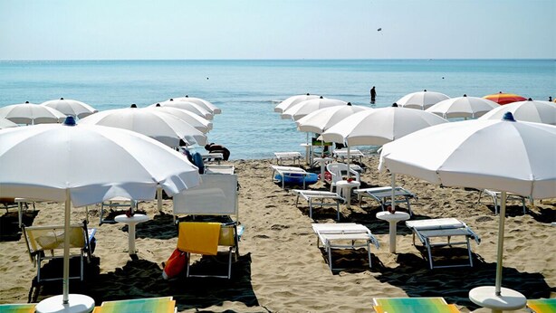 A beach with rows of white umbrellas and lounge chairs, some with towels. - Villaggio le Palatine (Bernalda)
