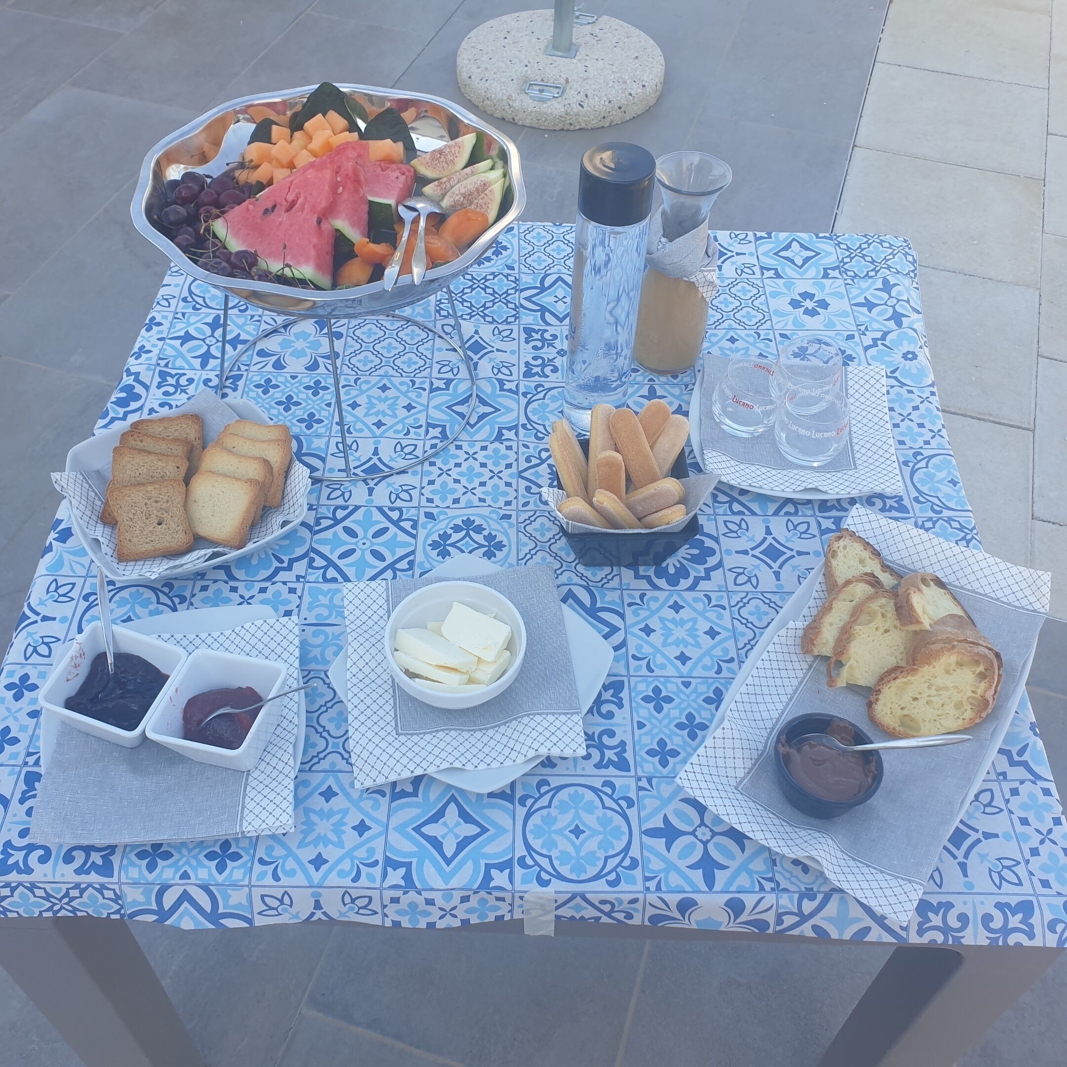 Une table dressée avec une variété d’aliments, notamment du pain, du fromage et des fruits, et ornée d’un motif décoratif en carreaux.