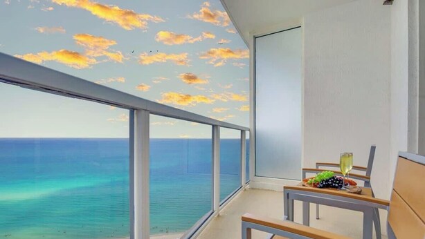 A balcony with a view of the ocean and a sunset sky. - Sunny Isles Bliss: Beachfront Retreat for 4 (Sunny Isles Beach)