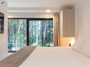 A bedroom with a large bed, a view of greenery through a window, and a wall-mounted light fixture.