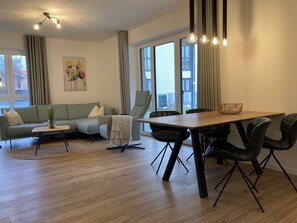 Een moderne woonkamer met een bank, een salontafel, een eettafel met stoelen en grote ramen.