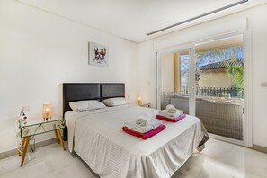 A hotel room with a large bed, a glass table, a nightstand, two bedside lamps, and a balcony with a view. - Vista Real 1 bedroom apartment (Nueva andalucia)