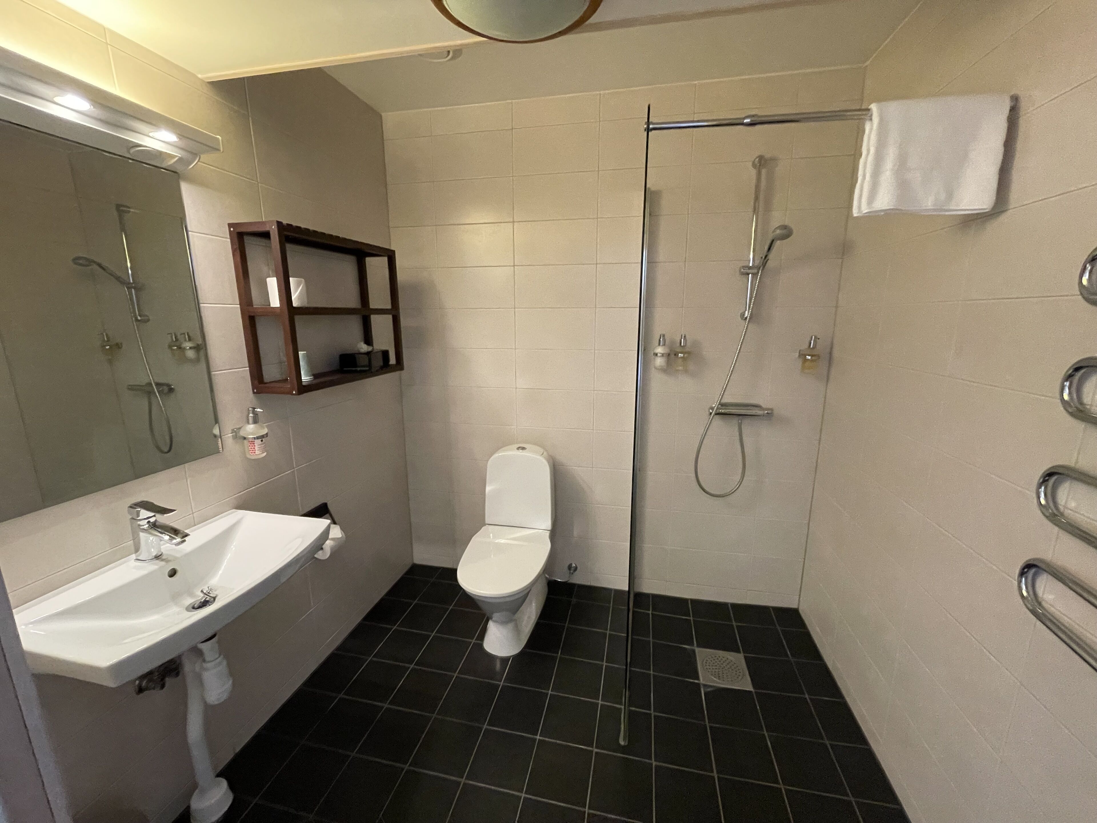 Un baño con lavamanos, inodoro y ducha blancos. Hay un espejo sobre el lavamanos y un perchero en la pared.