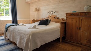 A neatly made bed with a white comforter, a dark pillow, and a rolled towel. There is a wooden nightstand with a lamp and a small blue object on top. A wooden cabinet with a decorative top is to the right of the bed. - Låsta Gårdshotell (Strängnäs)