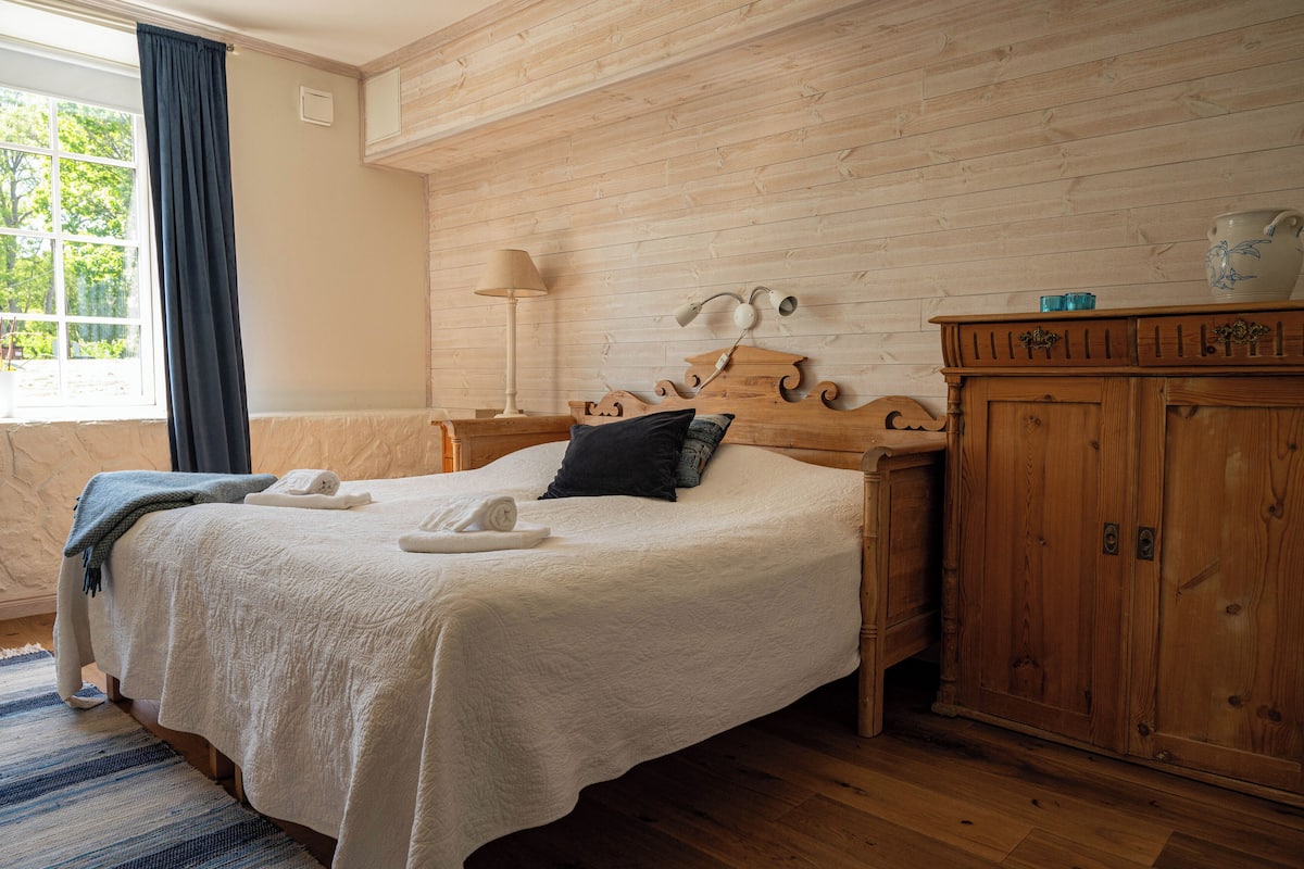 A neatly made bed with a white comforter, a dark pillow, and a rolled towel. There is a wooden nightstand with a lamp and a small blue object on top. A wooden cabinet with a decorative top is to the right of the bed.