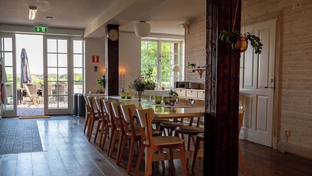 A dining area with wooden chairs and a table, a large window with a view, and a door with a hanging plant. - Låsta Gårdshotell (Strängnäs)