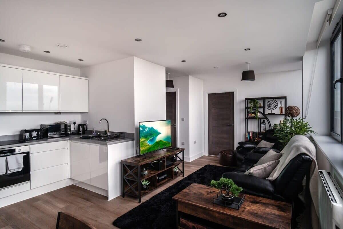 A modern kitchen with white cabinets, a black countertop, and a built-in oven. There is a wooden coffee table with a plant, a black sofa with cushions, and a TV on a wooden stand.