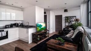 Una cocina moderna con armarios blancos, una encimera negra y un horno empotrado. Hay una mesa de centro de madera con una planta, un sofá negro con cojines y un televisor sobre un mueble de madera.