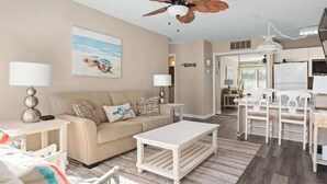 A living room with a beige sofa, white coffee table, and a ceiling fan. There is a painting on the wall and a kitchen area in the background. - Waterfront Ocean Villa! (Ocean City)
