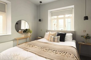 Een slaapkamer met een groot bed, een ronde spiegel, een raam met jaloezieën en een nachtkastje met een lamp.