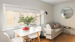 Een moderne woonkamer met een witte eettafel, een grijze bank en een groot raam met zonwering.