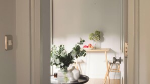 A room with a white wall, a small wooden table with a bowl of fruit, a vase with green plants, and a chair.