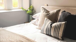 A neatly made bed with a white comforter and a beige throw blanket. There are several pillows in shades of beige and black. A small potted plant is visible on the windowsill. - Elegant King Suite · Behind Thermae Spa (Bath and North East Somerset)