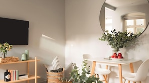 A modern dining area with a white table, chairs, and a mirror reflecting a window. There are baskets on shelves, a vase with flowers, and a bottle on the table.