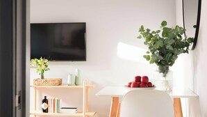 A modern living room with a flat-screen TV mounted on the wall, a white table with a plate of red apples, a vase with green foliage, and a bookshelf with books and decorative items. - Elegant King Suite · Behind Thermae Spa (Bath and North East Somerset)
