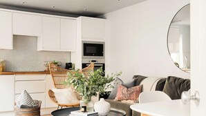 A modern kitchen with white cabinets, a microwave, and a built-in oven. There is a wicker chair and a white table with a vase of green plants. - Open-Plan Suite for 4 · Steps from Roman Baths (Bath and North East Somerset)