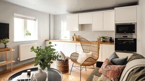 A modern kitchen with white cabinets, a built-in oven, and a microwave. There is a wicker chair and a sofa with cushions. A large window provides natural light. - Open-Plan Suite for 4 · Steps from Roman Baths (Bath and North East Somerset)