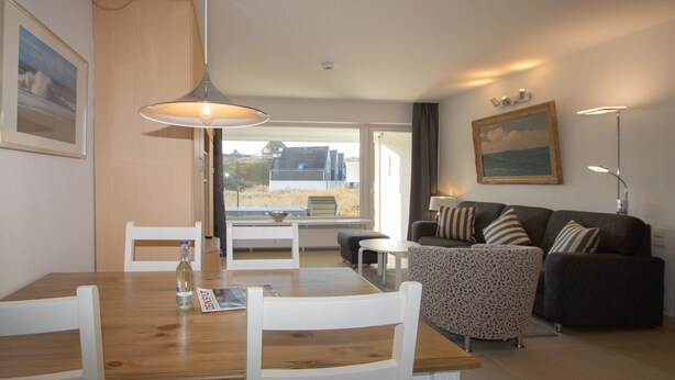 Uma sala de estar moderna com uma mesa de jantar de madeira, cadeiras brancas, um banquinho estampado, um sofá marrom escuro com almofadas listradas e um quadro na parede.