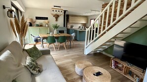 A modern living room with a sofa, dining table, and chairs. A staircase leads to an upper level. - Charming House Near Forest and Beach, Quiet Area, 3 Stars, WiFi, Garden, BBQ (Saint-Jean-de-Monts)