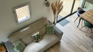 A modern living room with a sofa, a small window, a vase with pampas grass, and a dining area with a table and chairs. - Charming House Near Forest and Beach, Quiet Area, 3 Stars, WiFi, Garden, BBQ (Saint-Jean-de-Monts)