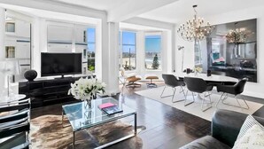 A modern living room with a large TV, a glass coffee table, and a dining area with a chandelier.