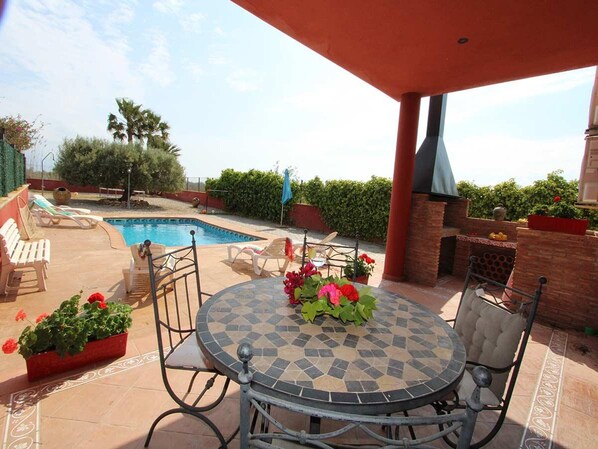 A poolside patio with a round table, chairs, and a barbecue grill.