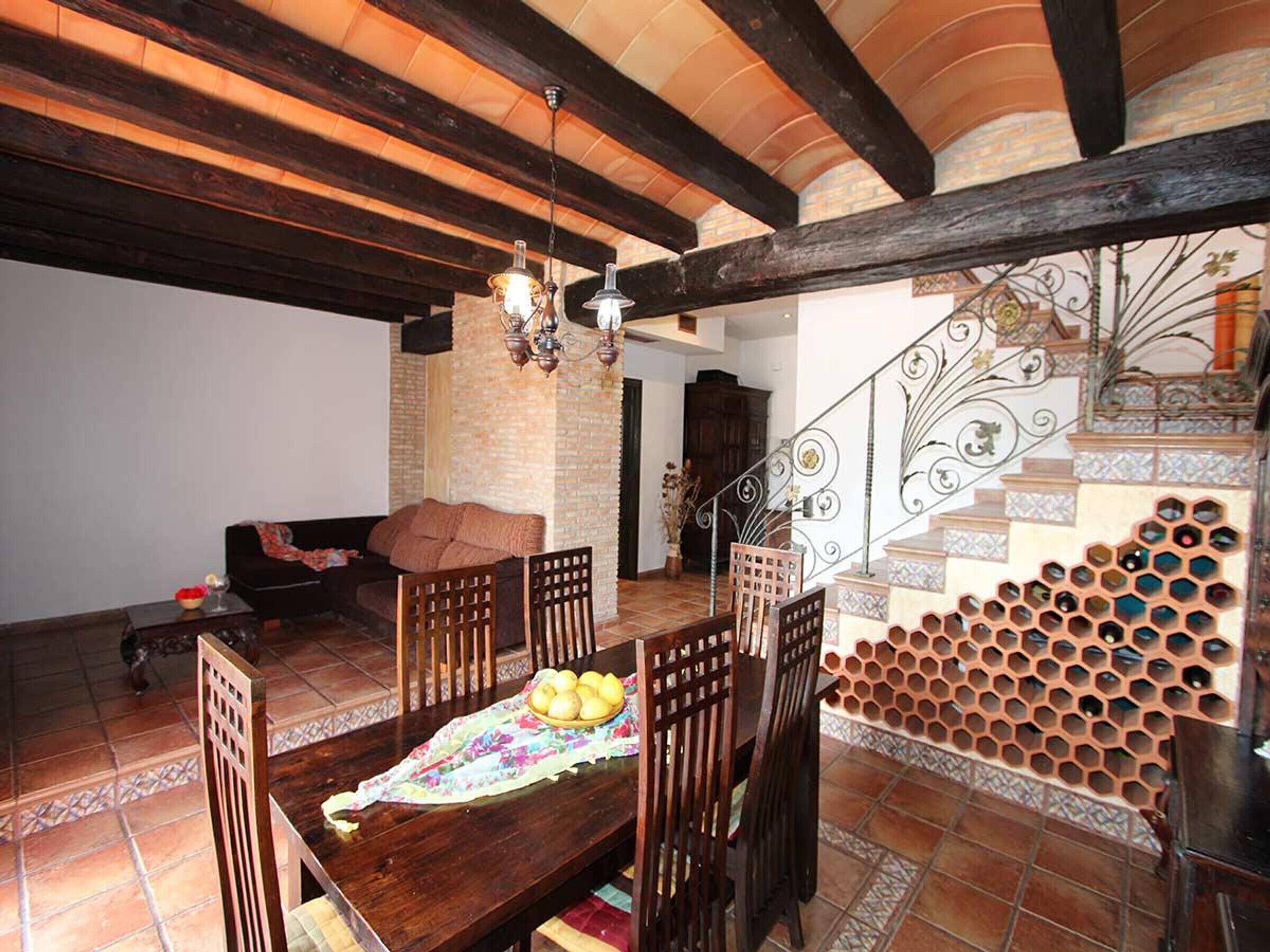 A dining area with a wooden table and chairs, a chandelier, and a staircase with a decorative metal railing.