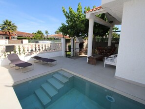 Uma piscina com escadas de acesso, cercada por espreguiçadeiras e uma área de estar coberta.